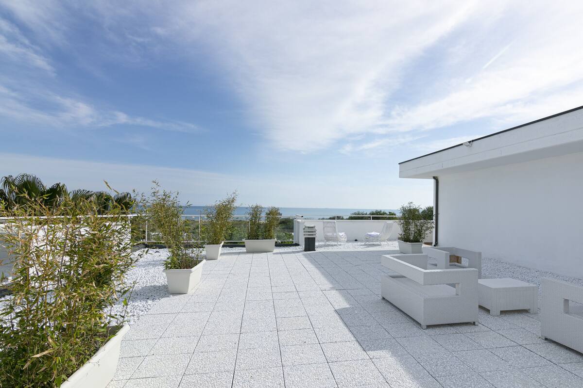 A rooftop patio with white wicker furniture, potted plants, and a view of the sea.