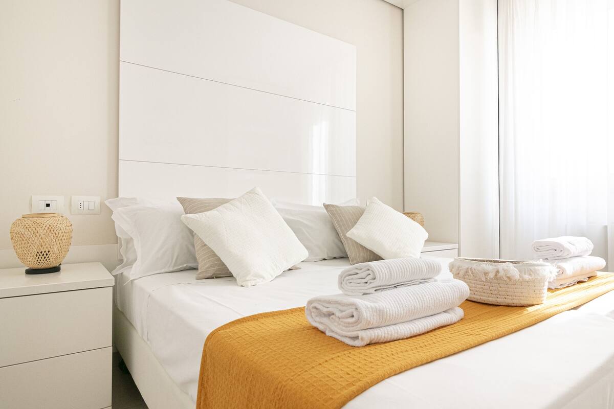 A hotel room with a neatly made bed, a bedside table with a lamp, and a window with curtains.