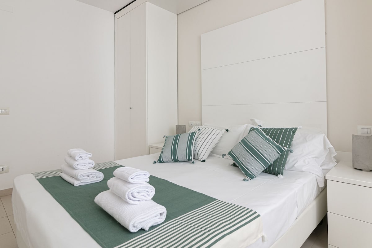 A hotel room with a neatly made bed, a green throw blanket, and several pillows. There is a nightstand with a lamp and a power outlet.