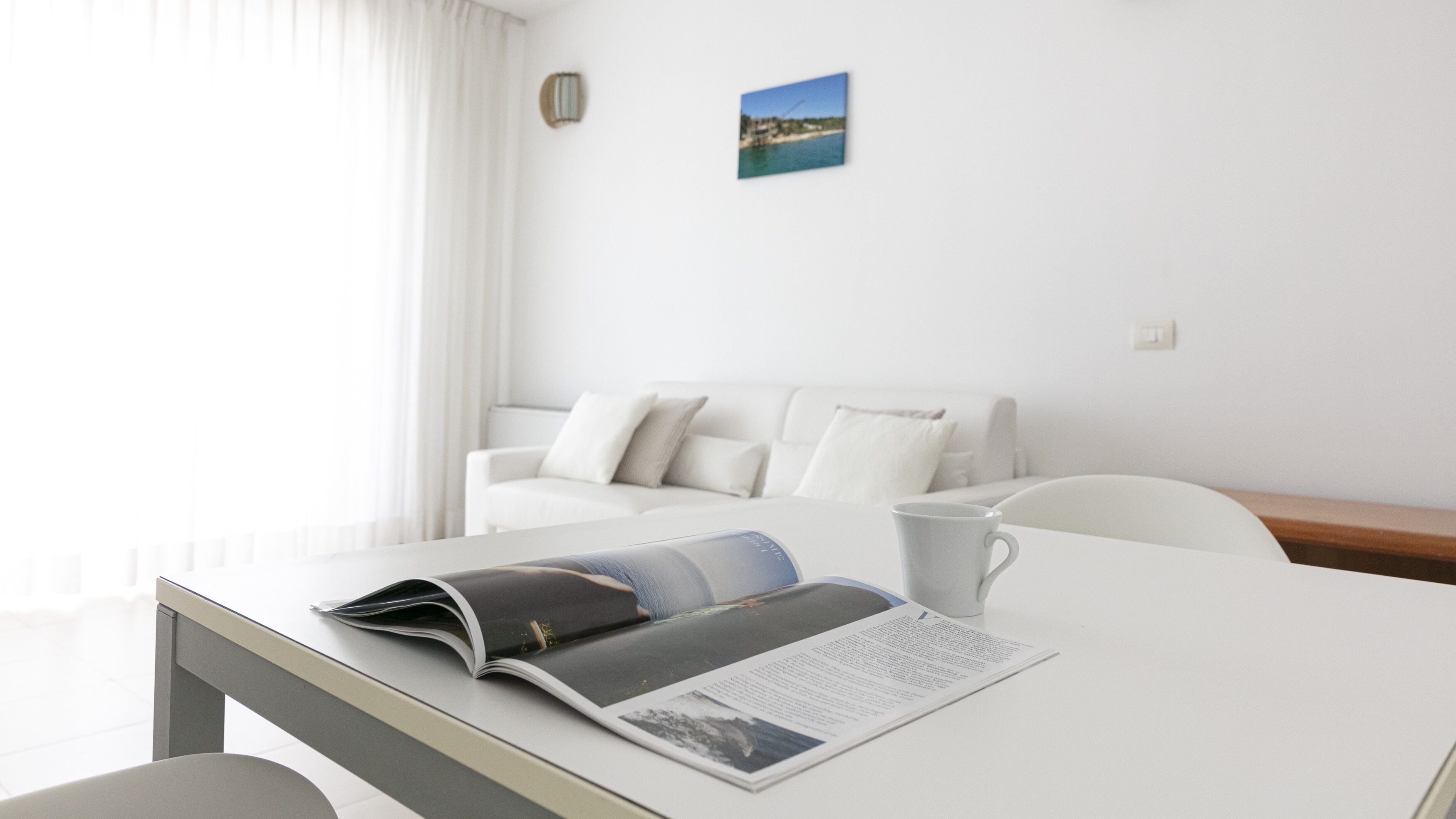 A modern, minimalist hotel room with a white color scheme, featuring a neatly made bed, a small table with a magazine and a cup, and a view of the sea through the window.