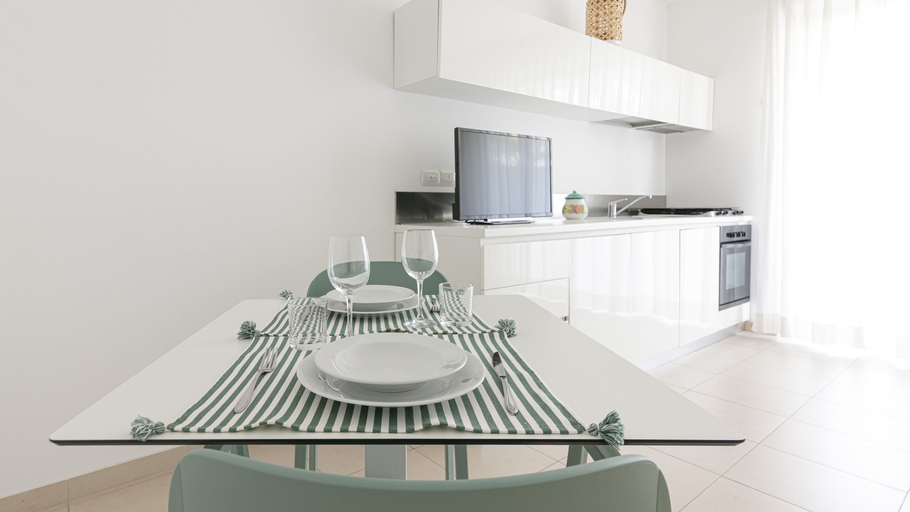 Une cuisine moderne avec des meubles blancs, une table à manger agrémentée de sets de table verts et blancs à rayures, et un téléviseur placé au-dessus du comptoir.
