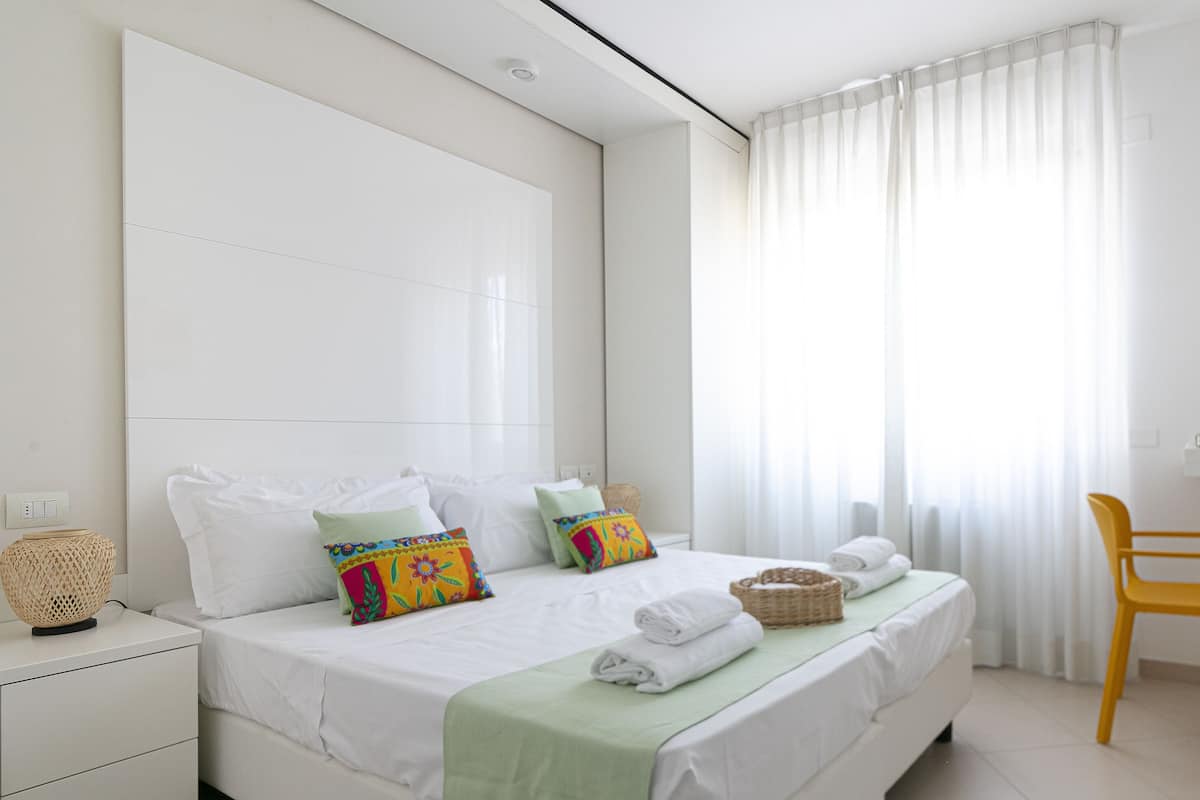A modern bedroom with a large bed, white bedding, colorful pillows, a wicker basket, and a yellow chair.