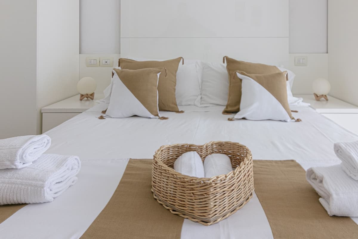 A hotel room with a neatly made bed, two nightstands with lamps, a wicker basket with towels, and a brown and white striped area rug.