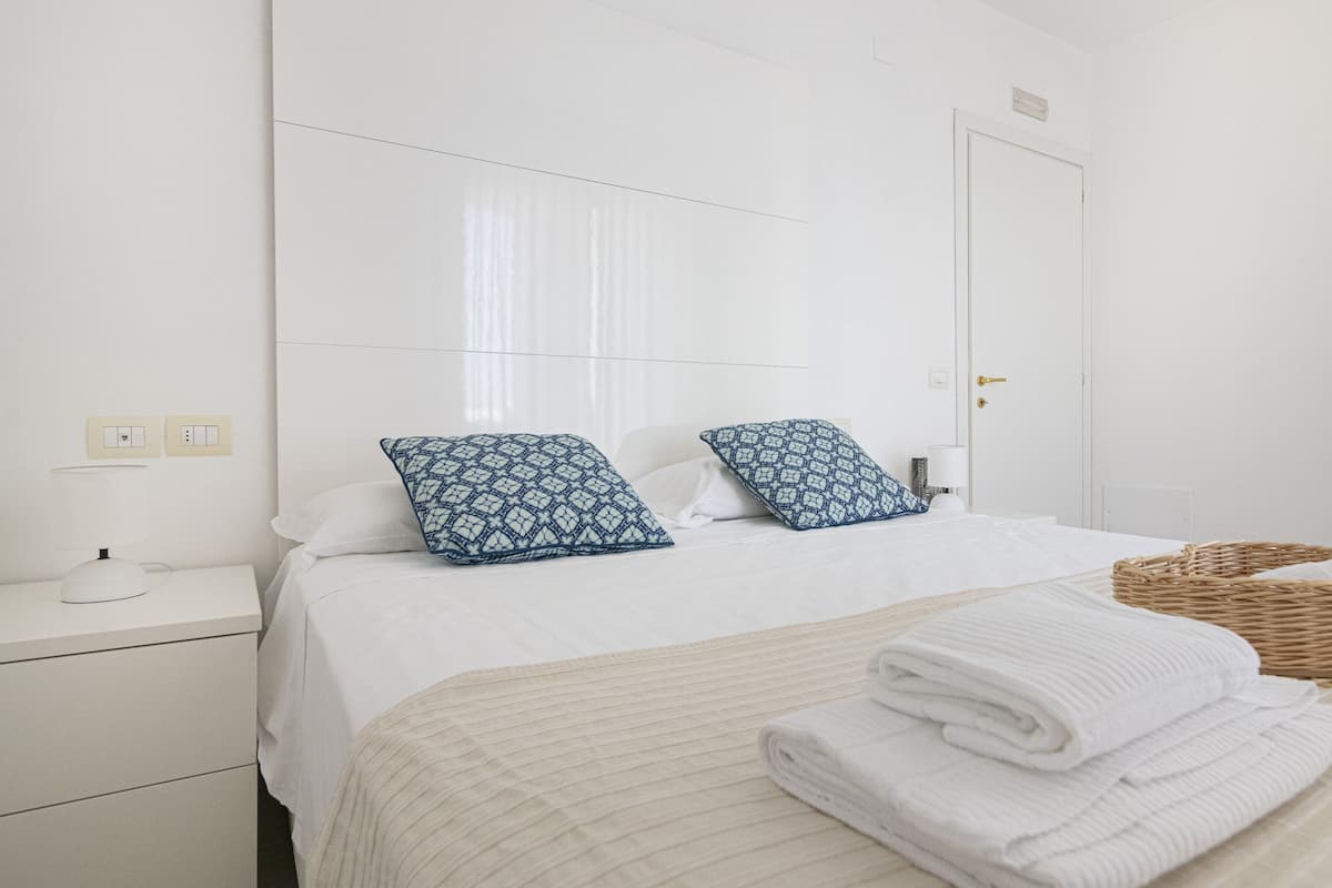 A hotel room with a bed, two pillows, a bedside table with a lamp, a basket of towels, and a door.