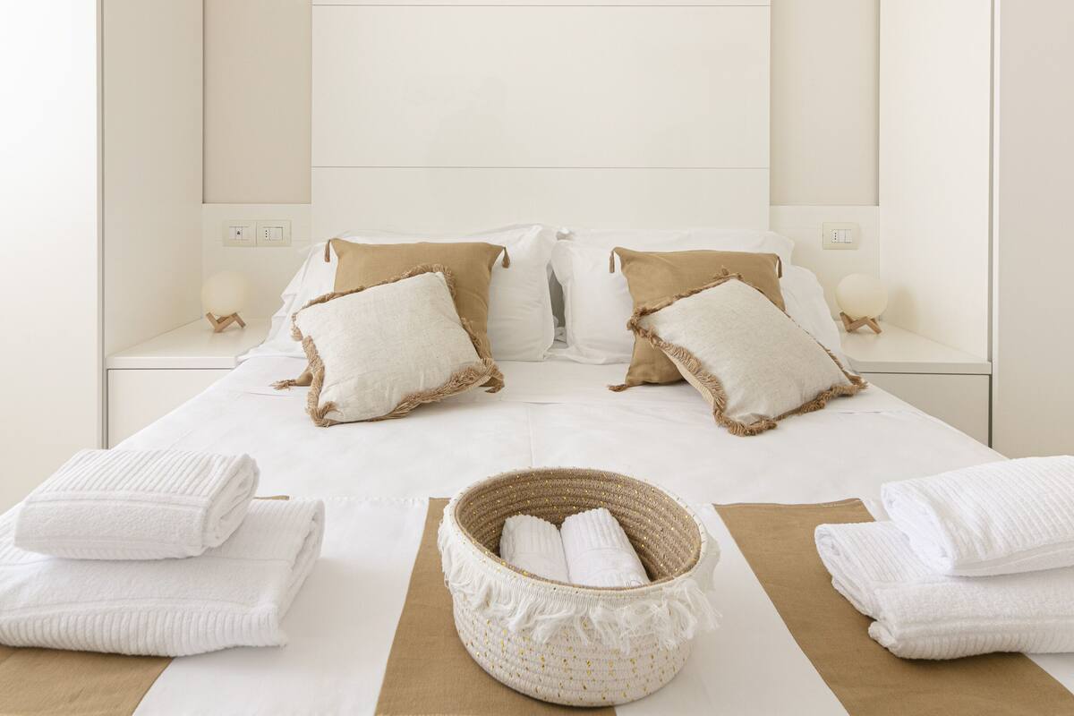A neatly made bed with white linens, two bedside tables with lamps, and a woven basket with towels.