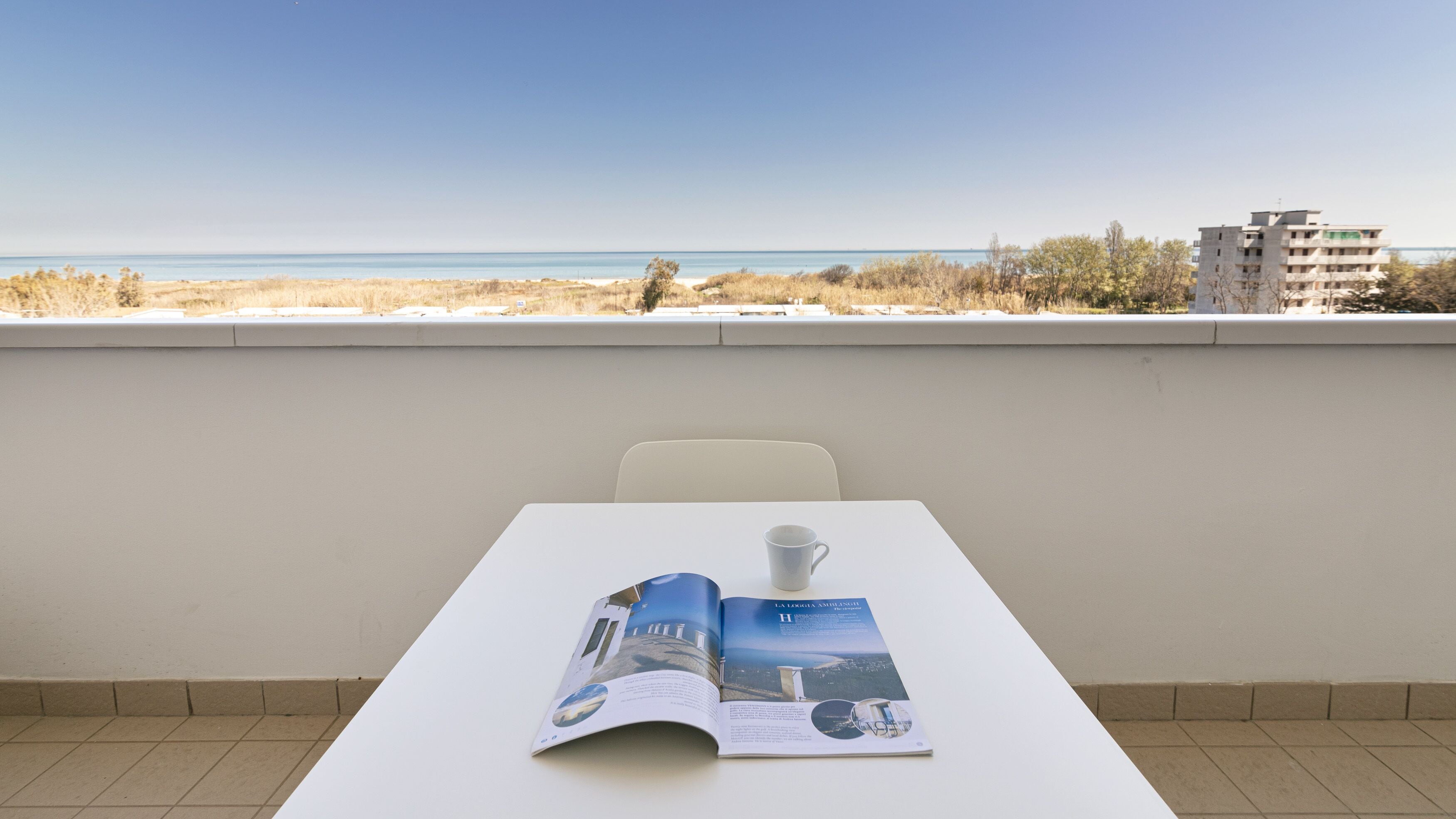 A table with a magazine and a cup, overlooking a beach and buildings.