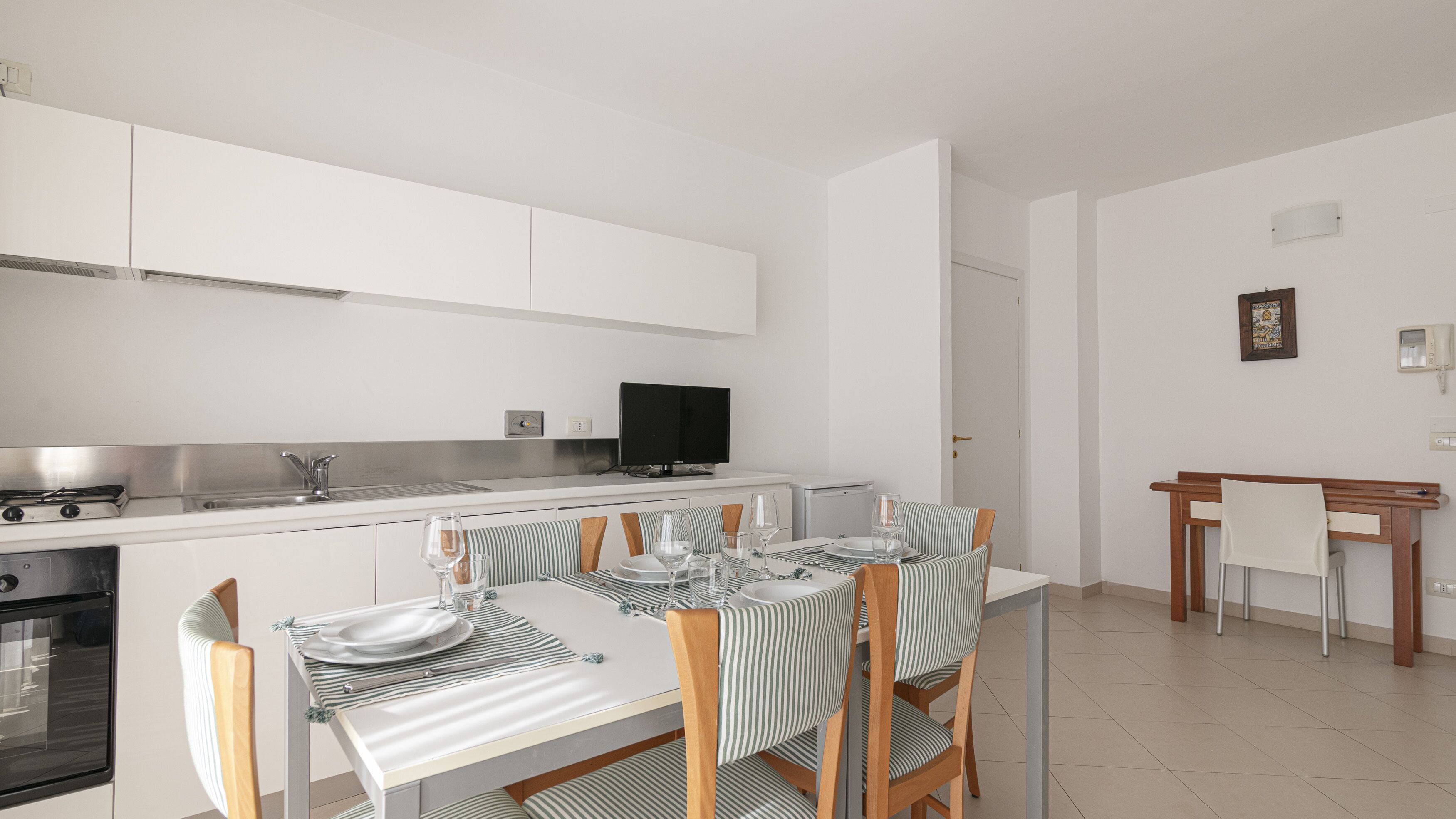 Una cucina moderna con mobili bianchi, piano cottura integrato e zona pranzo con sedie a righe e tavolo apparecchiato per due.