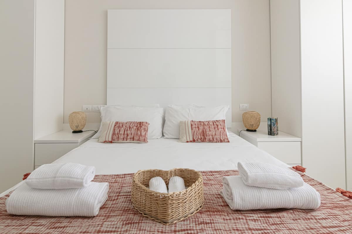 A hotel room with a large bed, two bedside tables, a bedside lamp, and a woven basket with towels.