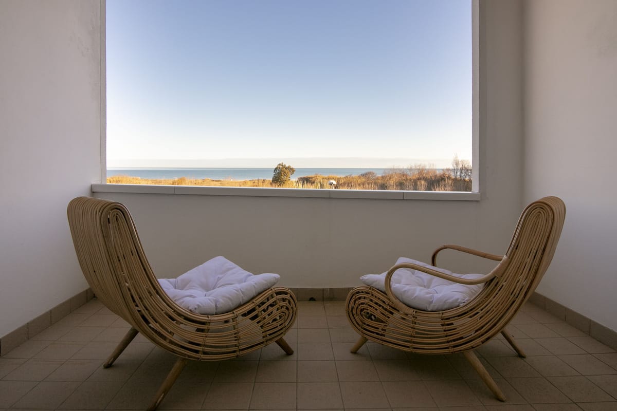 Two wicker chairs with white cushions facing a large window overlooking a beach.