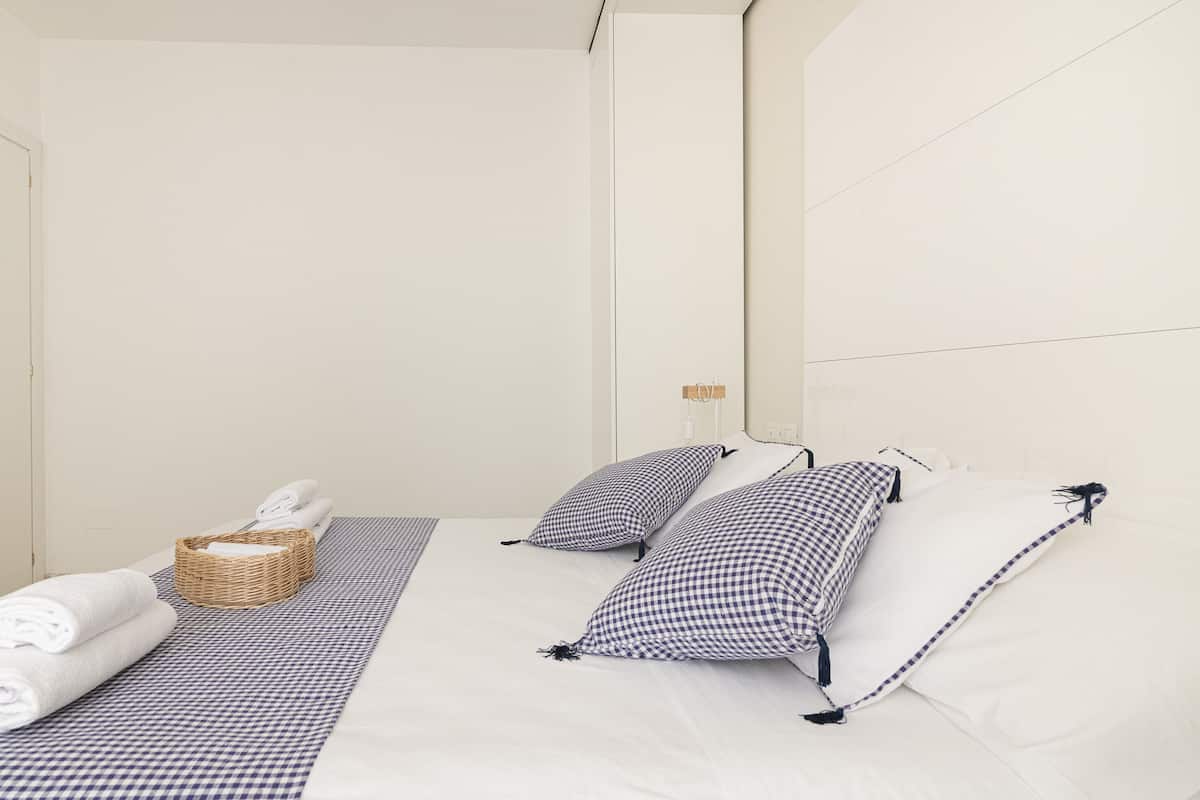 A hotel room with a bed, pillows, a basket with towels, and a checkered mat.