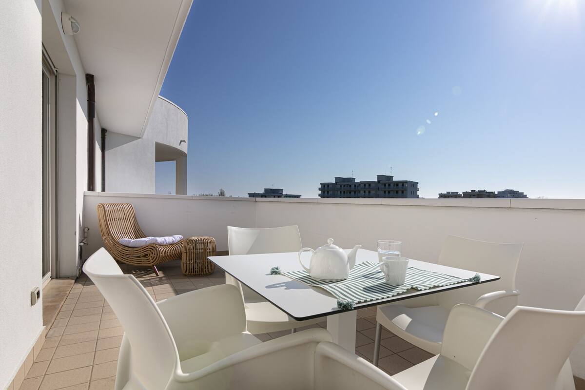 A balcony with a table set for two, white chairs, a wicker chair, and a view of buildings in the distance.