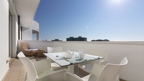 Un balcon avec une table dressée pour deux personnes, des chaises blanches, un fauteuil en osier et une vue sur les bâtiments au loin.