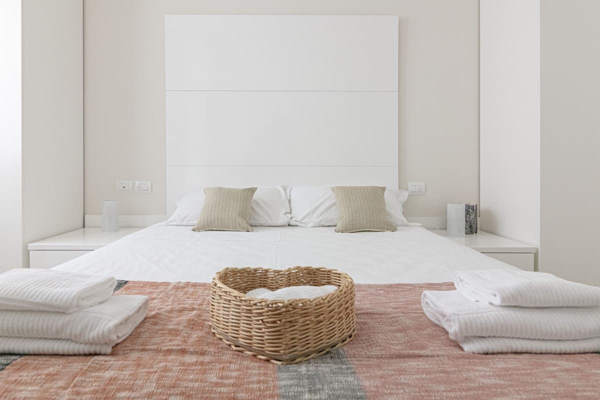 A minimalist bedroom with a large bed, two bedside tables, a wicker basket, and folded towels.