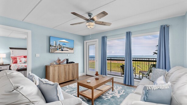 A living room with a ceiling fan, a wooden coffee table, a sofa, and a view of a balcony with a railing. - Stunning Remodeled Condo on the Beach! Resort Pools, Hot Tubs, Tennis Courts & More! Dog-Friendly! (South Padre Island)
