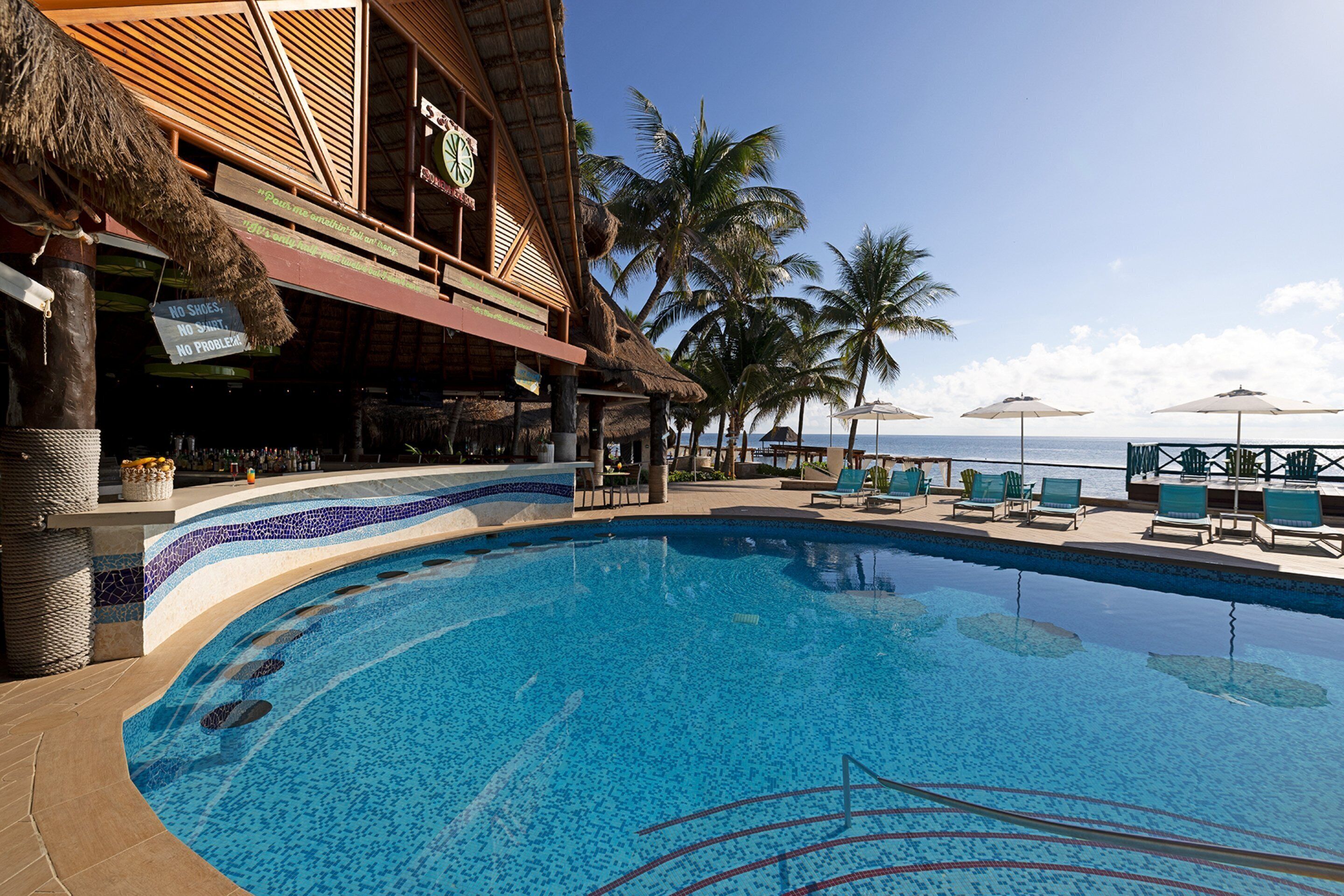 Un espace piscine avec des chaises longues et des parasols, un bar et des palmiers.