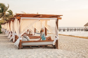 A row of wooden beach cabanas with white canopies, arranged along a sandy beach with a wooden pier in the background.