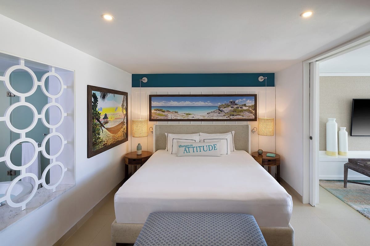 A hotel room with a bed, two wall-mounted lamps, a flat-screen TV, and two paintings.