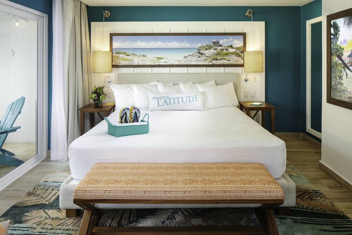 A neatly arranged hotel room with a large bed, a bench, a small table with a basket, and a view of a beach through a window.