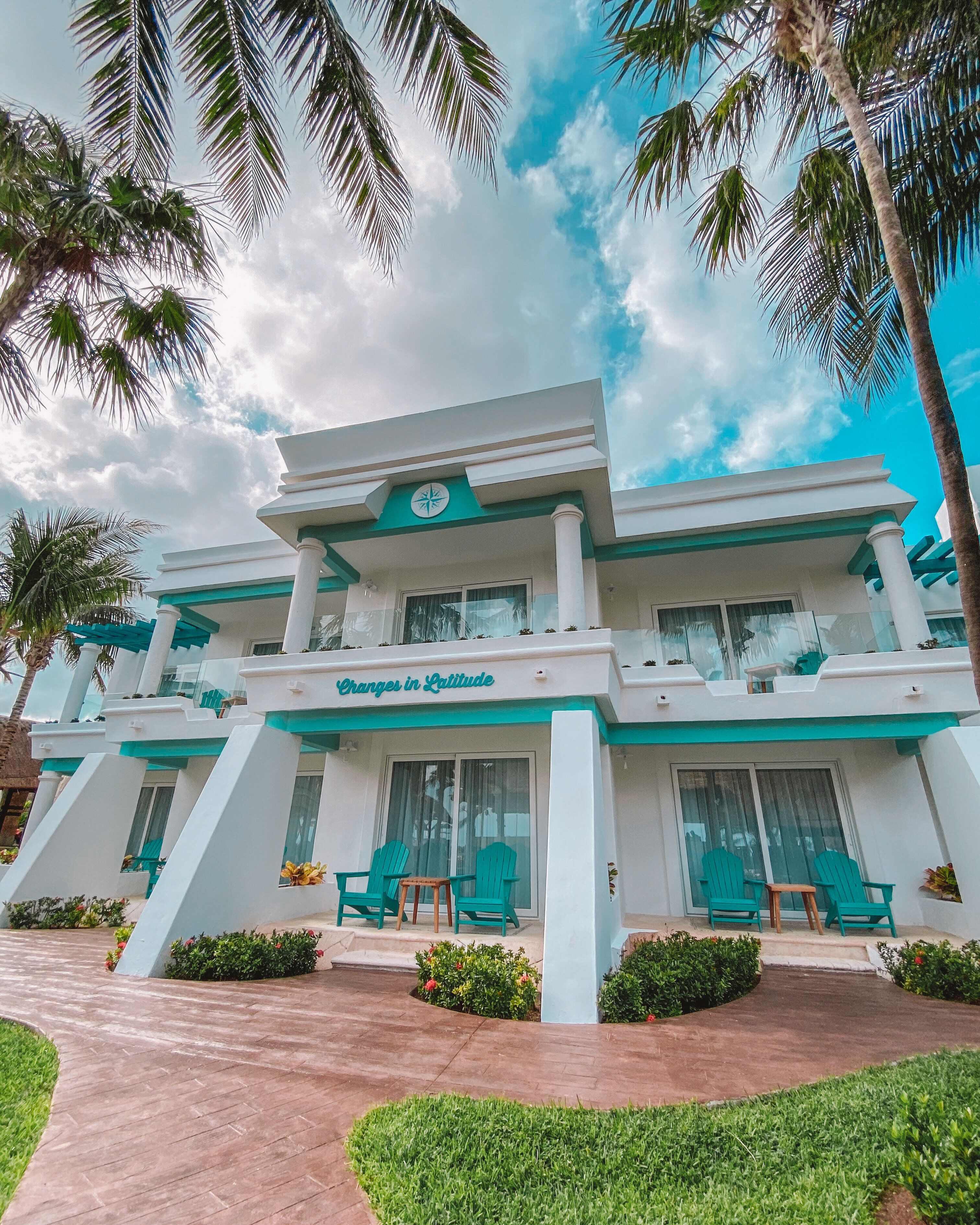 A two-story hotel with a modern design, featuring a prominent sign that reads "Oranges in Latitude." The building has a flat roof, white exterior walls, and a series of balconies with green chairs and small tables.