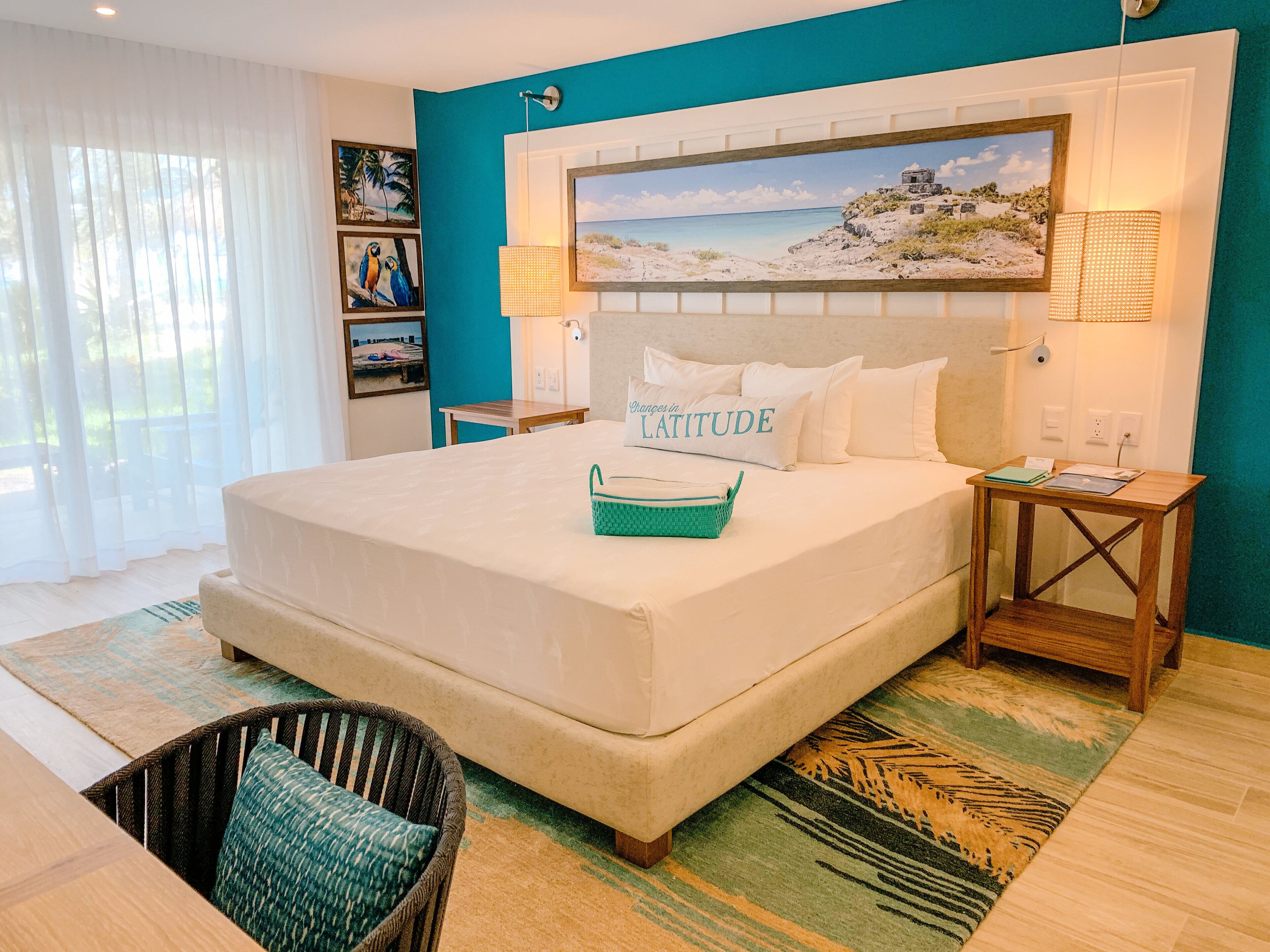 A neatly made bed with a pillow labeled "Changes in LATITUDE," a teal woven basket, and a wooden nightstand with a lamp.