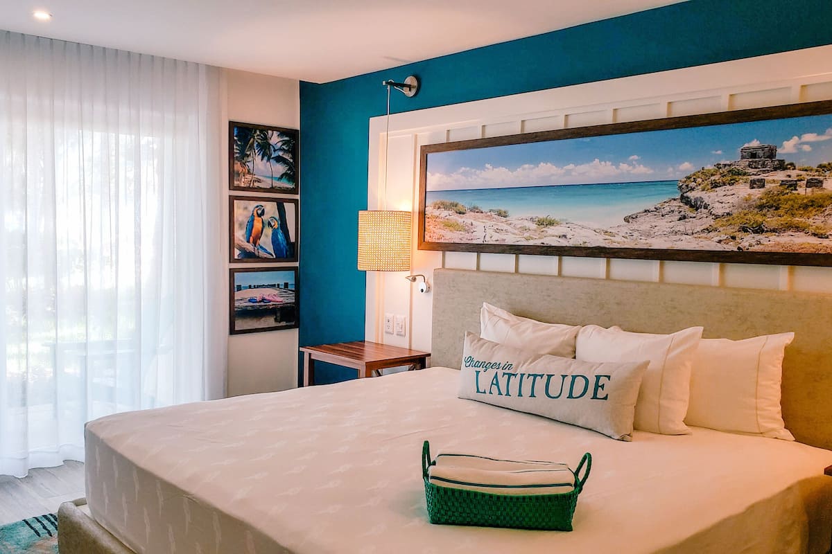 A hotel room with a large bed, a painting of a beach scene, a lamp, and a small table.