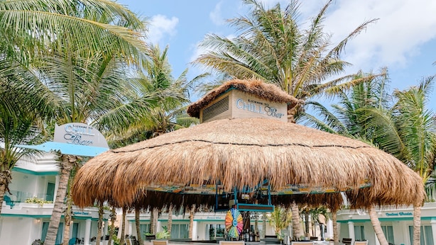 Un bar con techo de paja y zona de estar circular, rodeado de palmeras y una piscina.
