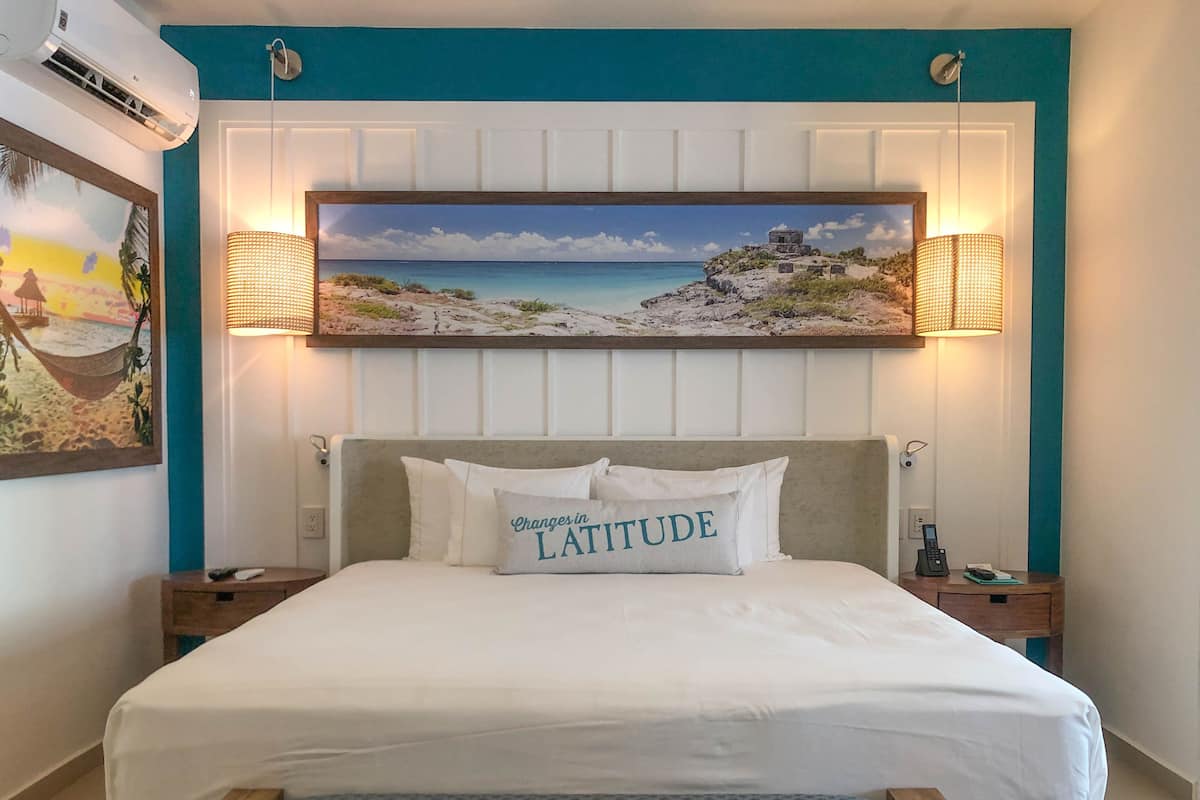 A hotel room with a bed, a painting of a beach scene, and two wall-mounted lamps.