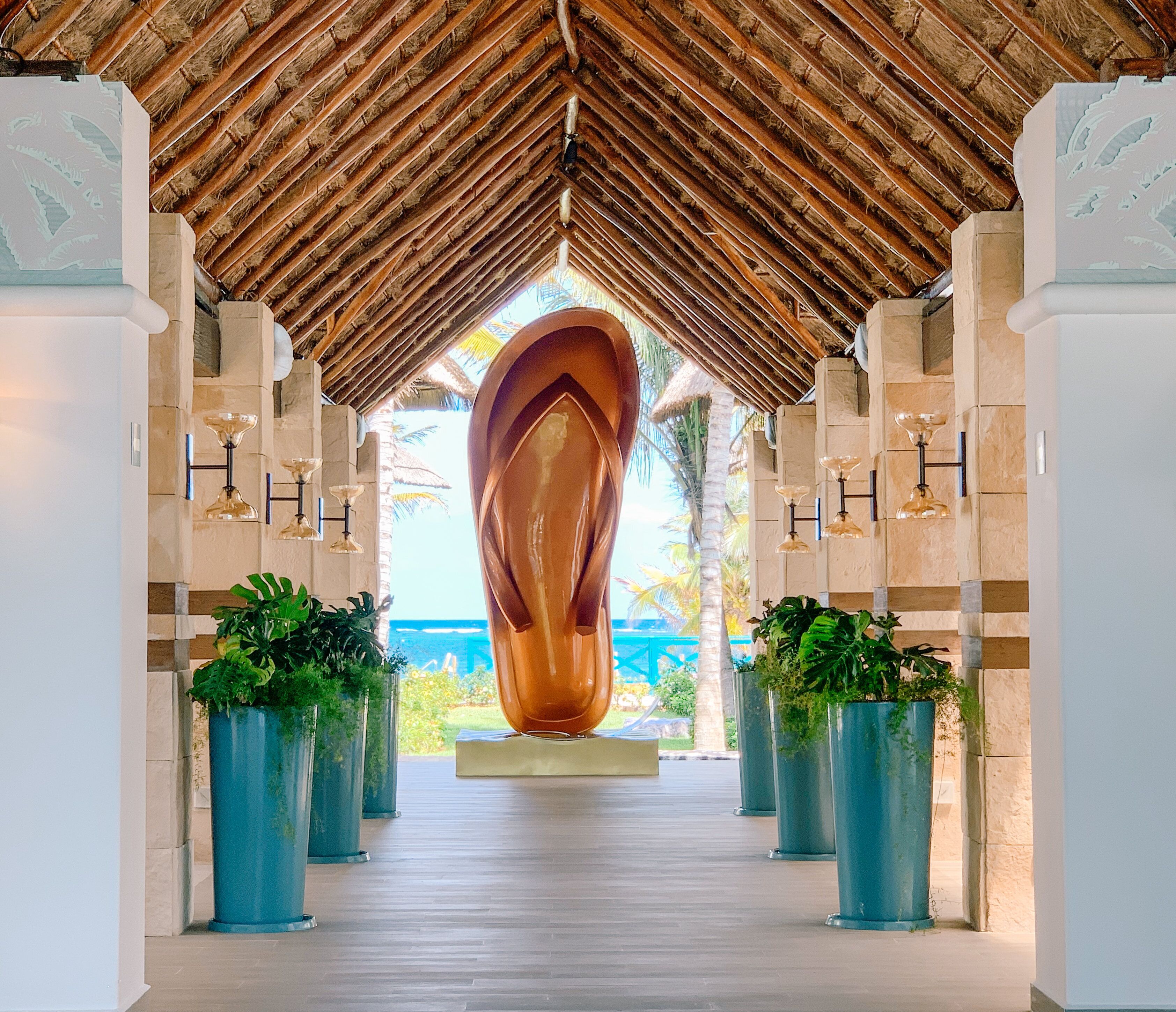 A covered walkway with a central sculpture, flanked by columns and potted plants, leading to a view of the sea.