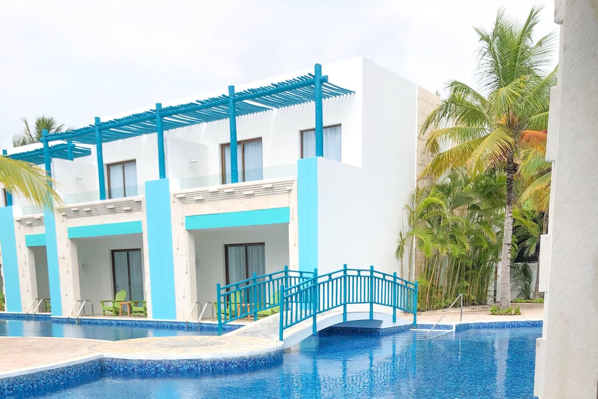 A pool area with a wooden table and chairs, a blue bridge, and a view of a building with blue and white walls.