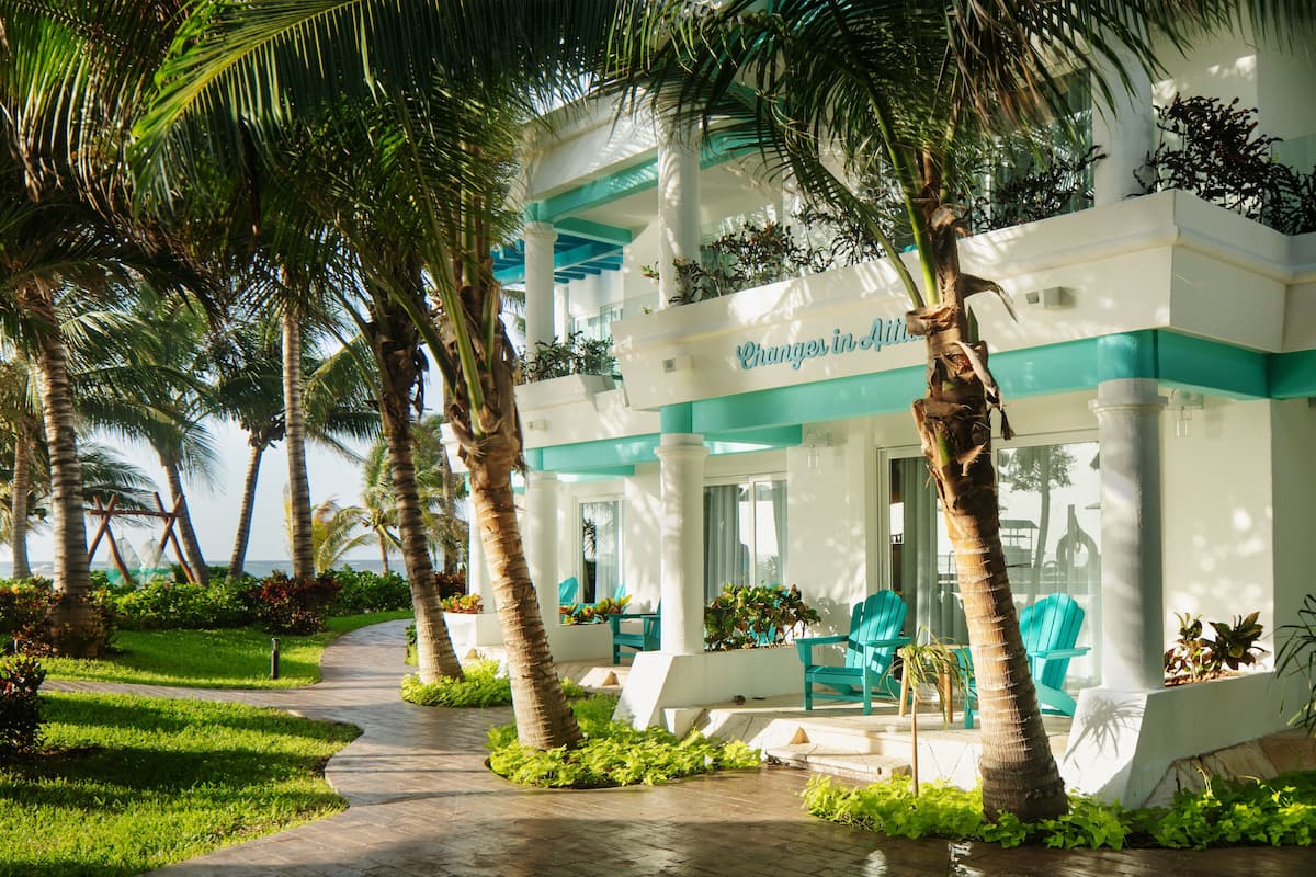 A resort with a pathway lined with palm trees, a pool area with blue chairs, and a sign reading "Changer in Aji."