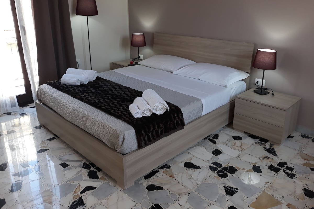 A neatly made bed with white linens and a dark throw blanket, two bedside lamps, a wooden nightstand, and a tiled floor.