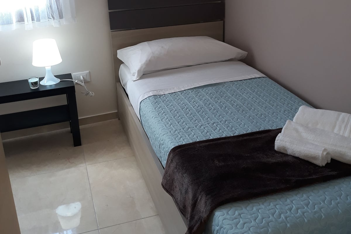 A small, neatly arranged bedroom with a single bed, a bedside table with a lamp, and a window with sheer curtains.