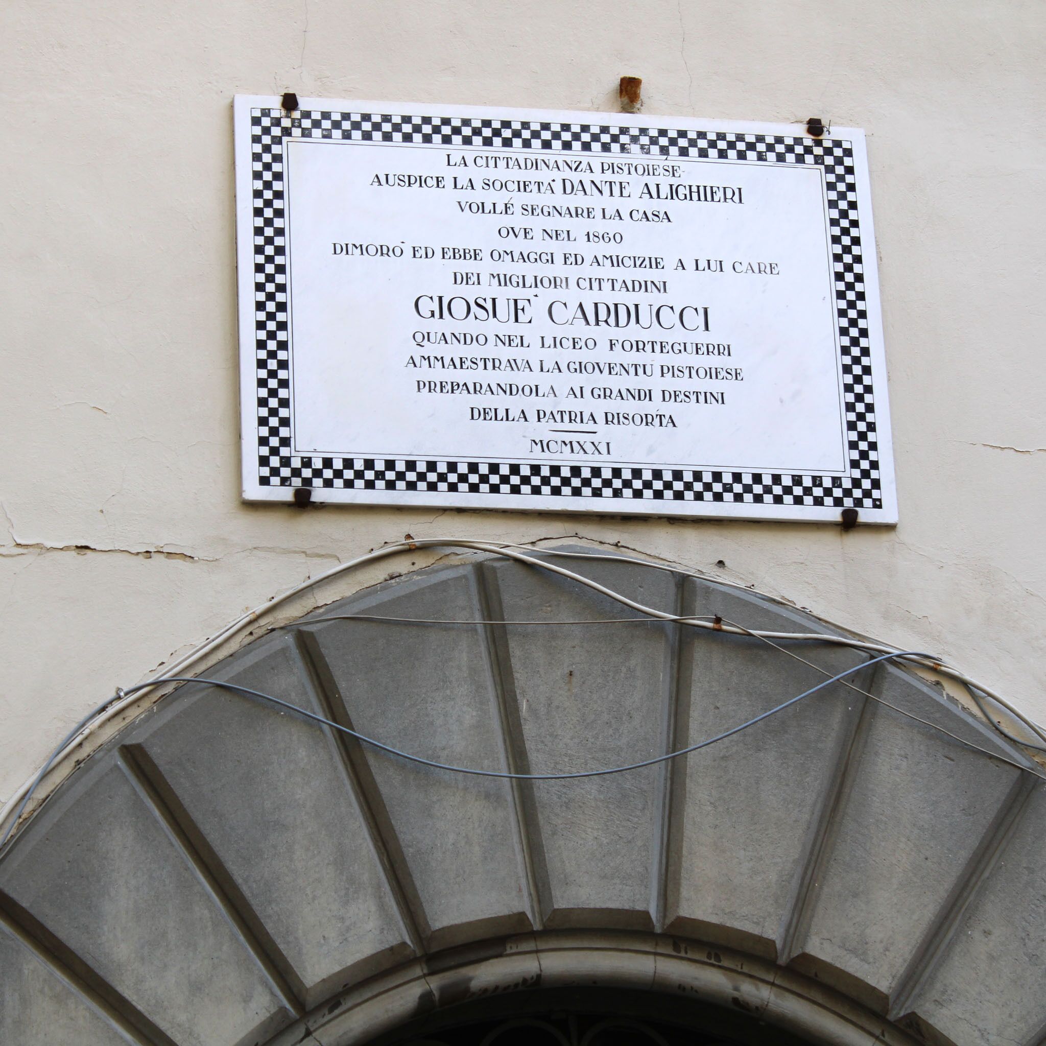 Une plaque avec du texte en italien, fixée à un mur dont le bord est décoré d’un motif à carreaux.