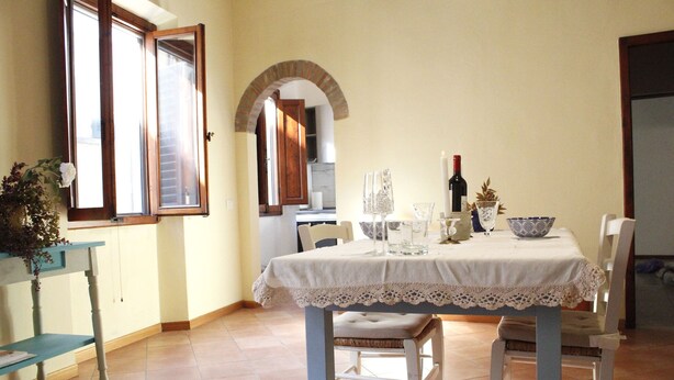 A dining area with a table set for two, a window with open shutters, and a view into the kitchen. - Carducci bright flat in historic centre (Pistoia)