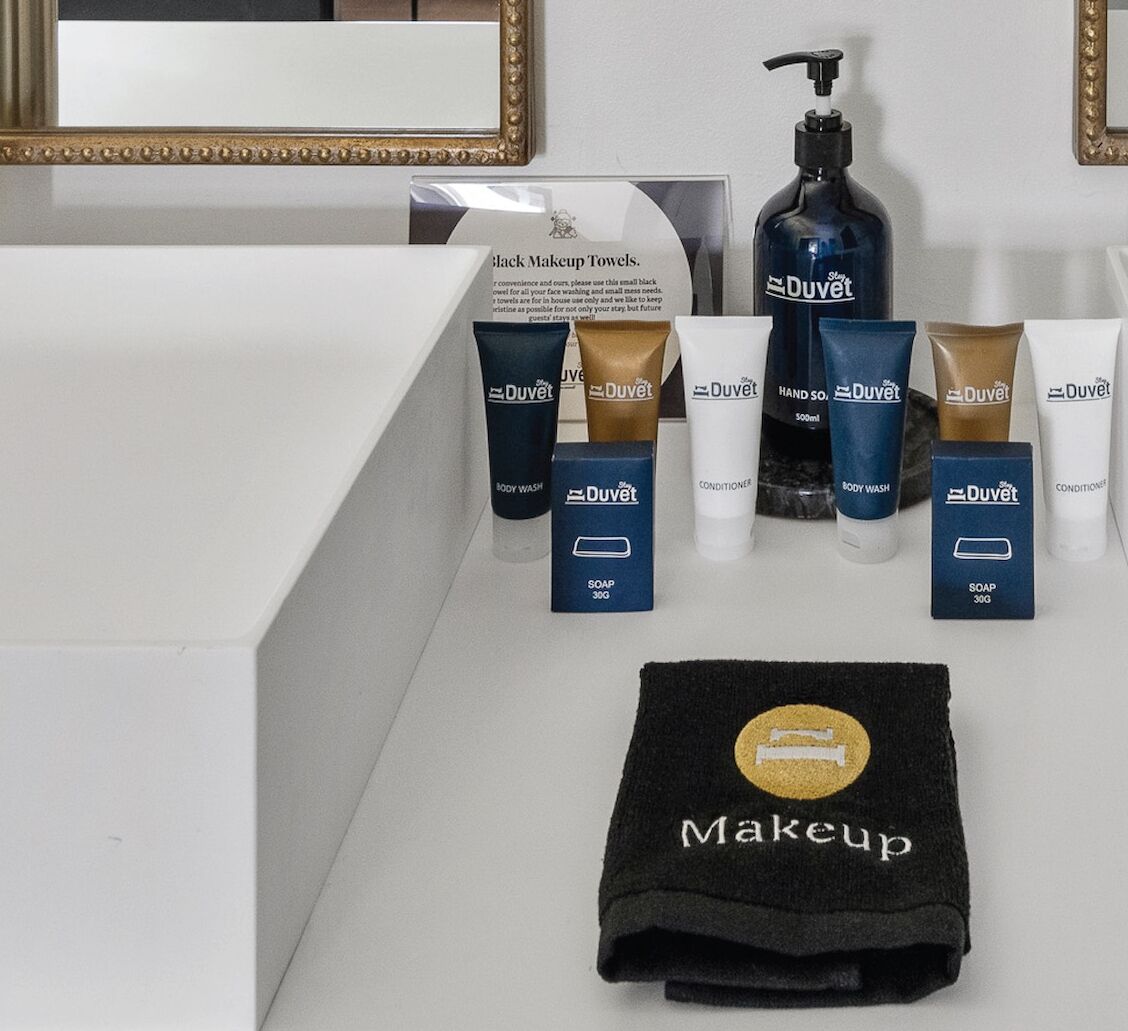 Un tocador de baño con una toalla negra para maquillaje, varios productos de crema y jabón DuVet, y un espejo con marco dorado.