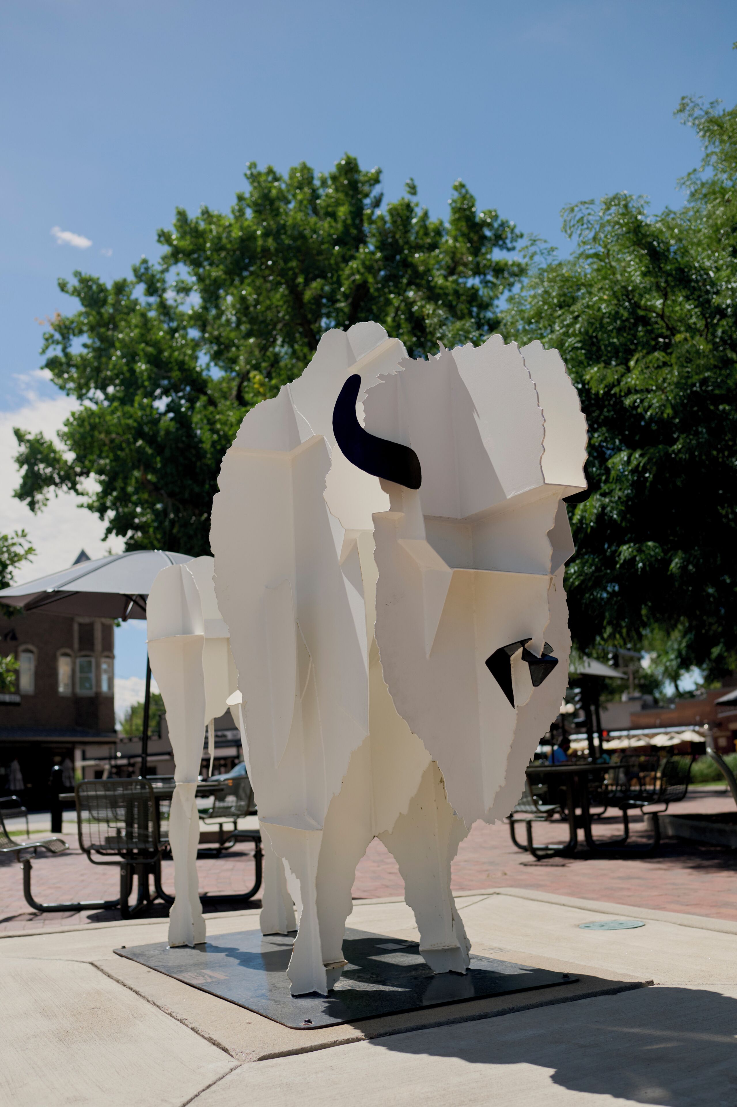 Una escultura abstracta blanca en un entorno exterior, con árboles y un edificio al fondo.