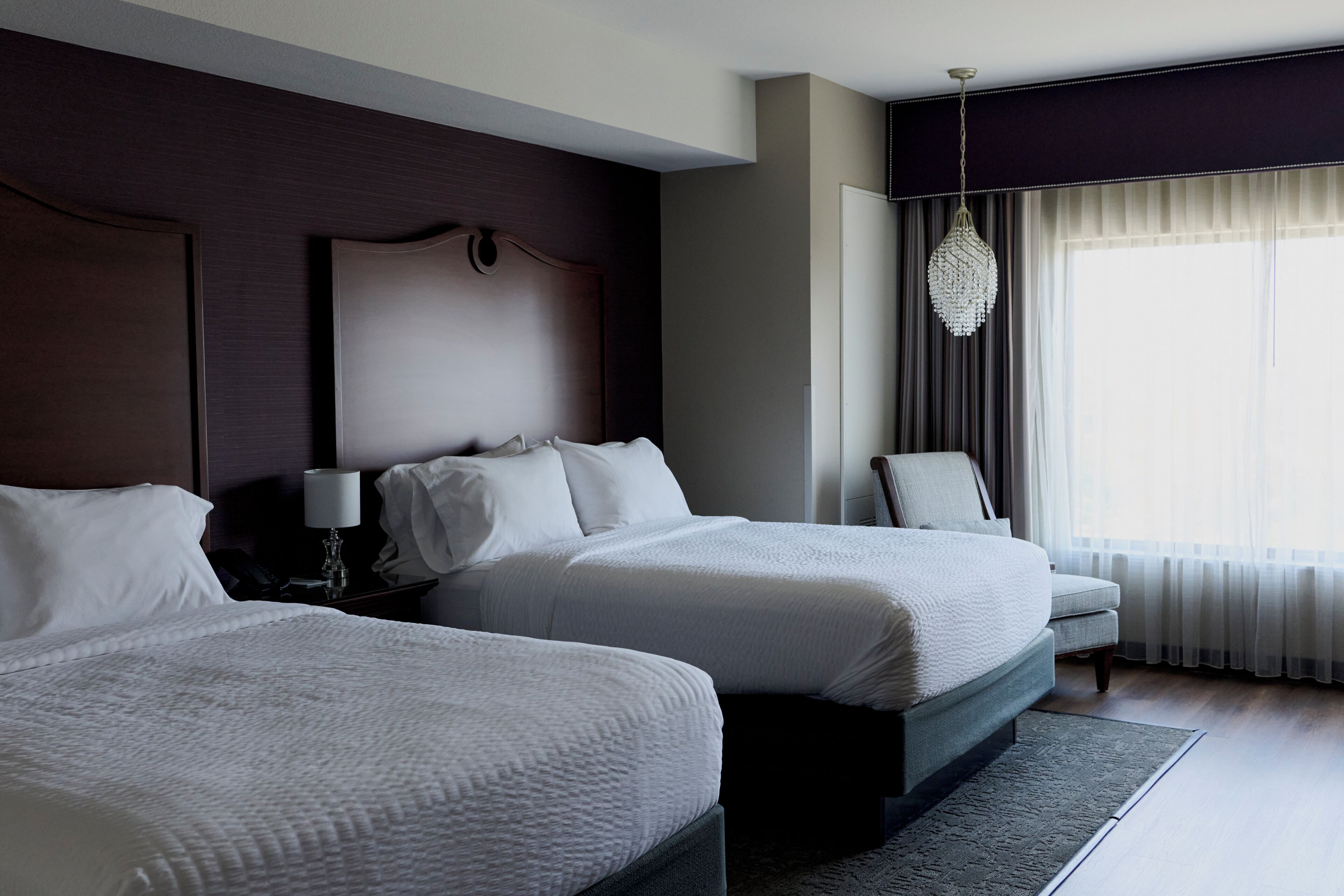 Quarto de hotel com duas camas, uma mesinha de cabeceira, uma luminária, uma cadeira e uma janela grande com cortinas.
