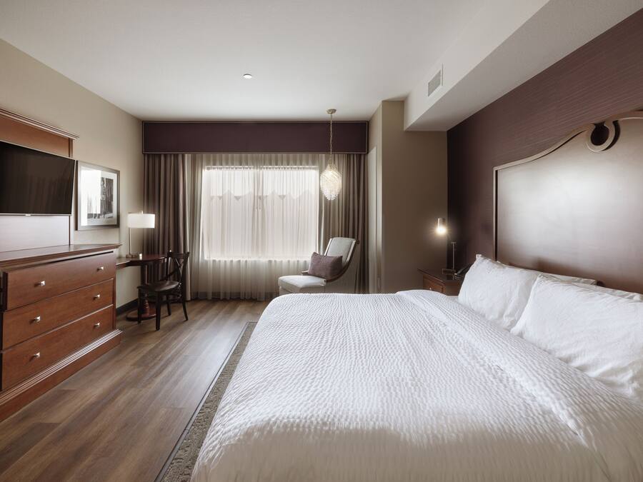 Habitación de hotel con una cama grande, un escritorio con silla, televisión y ventana con cortinas.