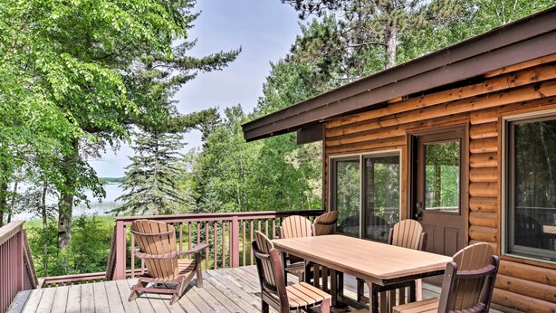 A wooden deck with a table and chairs, overlooking a forested area and a body of water. - Rustic Cozy Cabin on Island Lake w/ Fire Pit, Dock (Park Rapids)