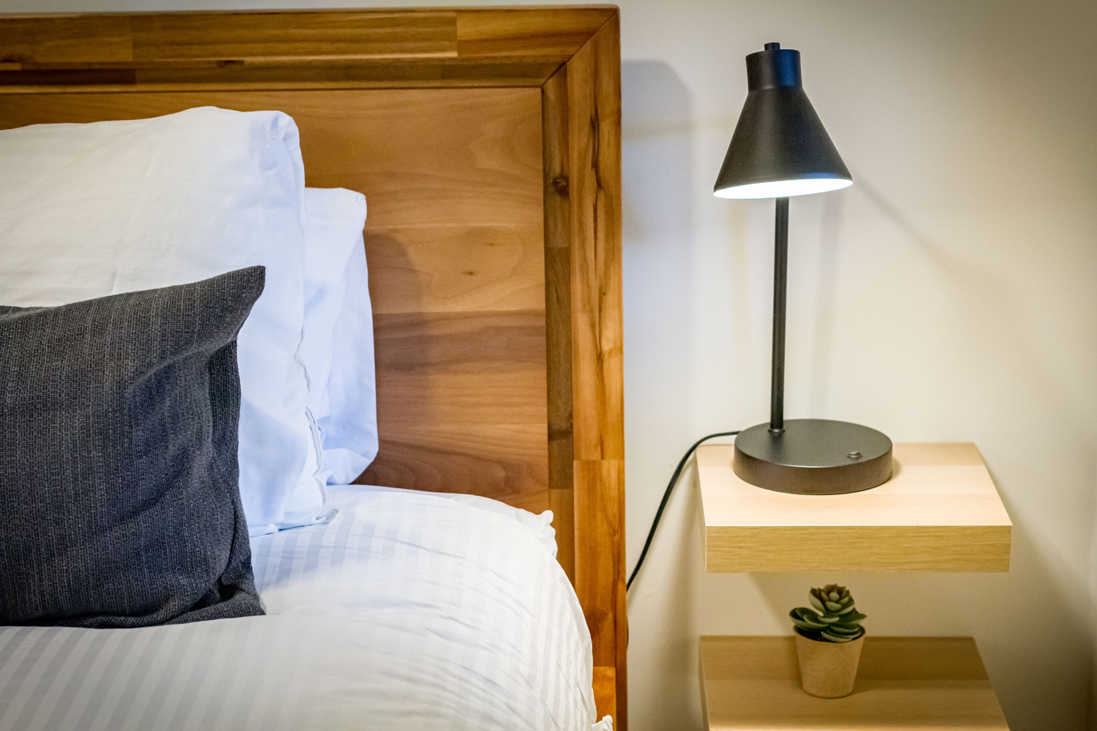 Ein ordentlich bezogenes Bett mit weißen und grauen Kissen, ein hölzernes Kopfteil und eine schwarze Schreibtischlampe auf einem Nachttisch aus Holz mit einem kleinen Topfblumen.