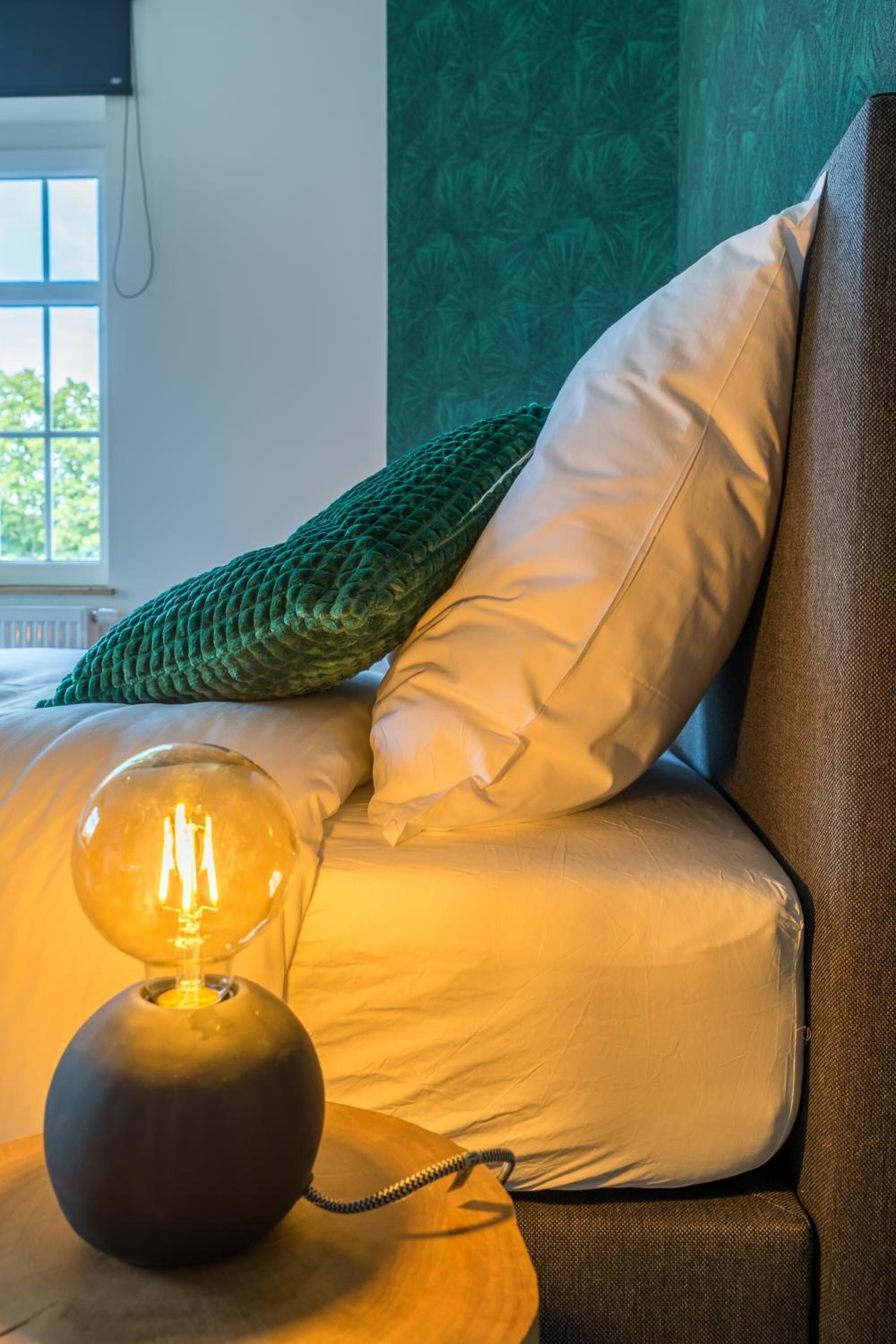 A cozy room with a bed, a green knitted pillow, a white pillow, and a table lamp.