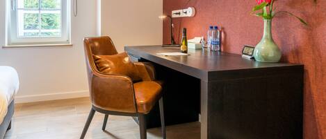 A hotel room with a desk, a brown leather chair, a vase with a flower, a television mounted on the wall, and a window with a view of greenery.