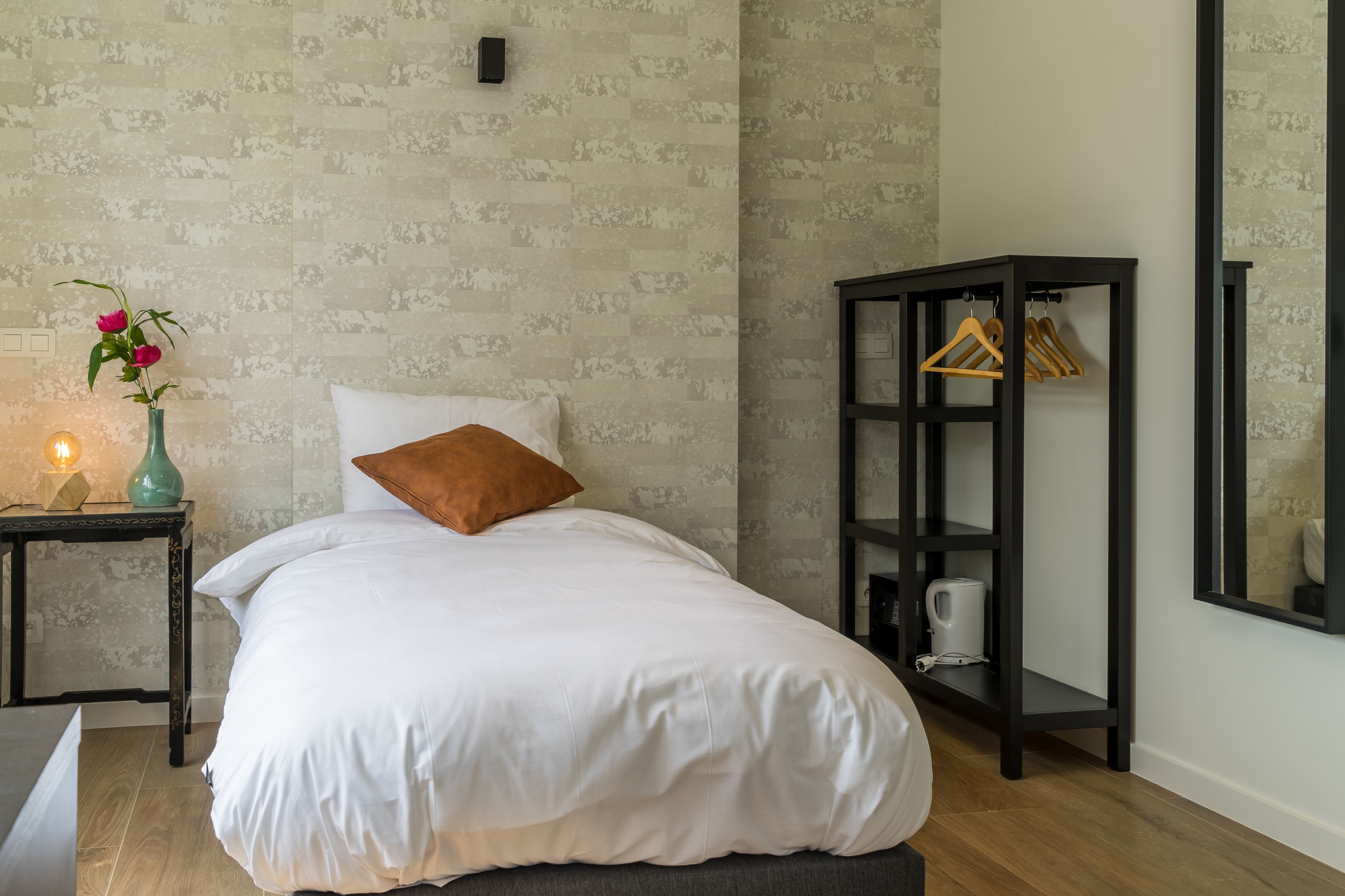 Una cama bien hecha con edredón blanco y almohada marrón, una mesita con un florero verde azulado y una vela encendida, y un estante negro con perchas de madera.