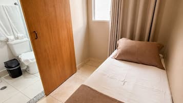 Un dormitorio con cama, ventana con cortinas, puerta de madera y baño a la vista al fondo.