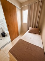 Un dormitorio con cama, ventana con cortinas, puerta de madera y baño a la vista al fondo.