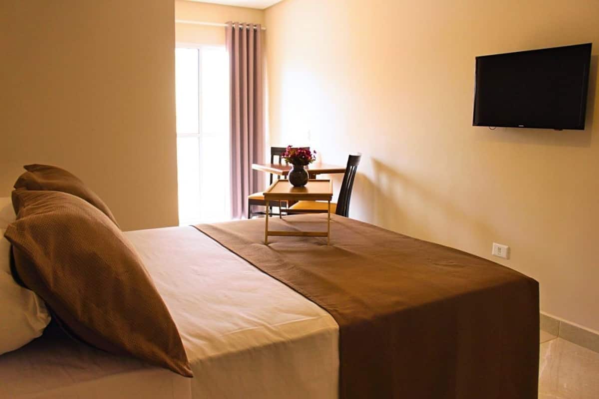 Une chambre d’hôtel avec un lit, une table et des chaises, un vase avec des fleurs et un téléviseur fixé au mur.