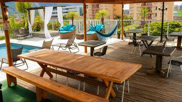 A covered outdoor area with wooden tables and benches, a pool, and lounge chairs.