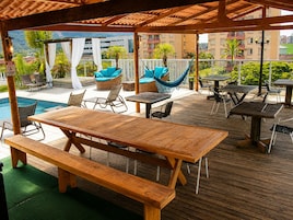 A covered outdoor area with wooden tables and benches, a pool, and lounge chairs.