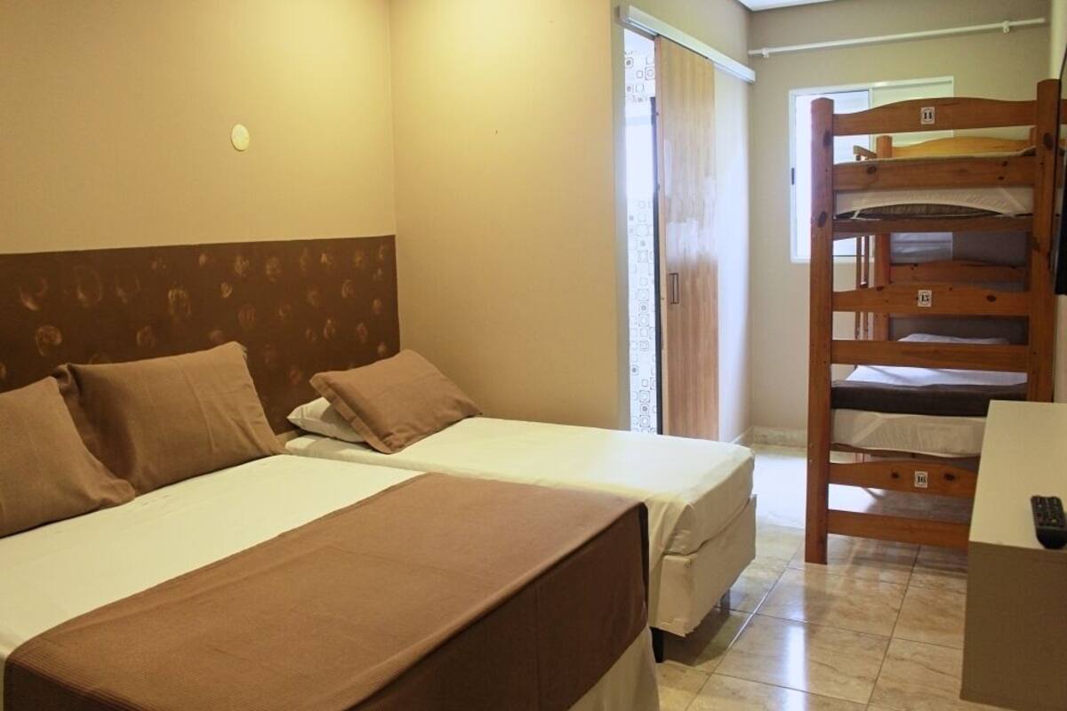 Une chambre d’hôtel avec deux lits, un lit superposé en bois, une fenêtre avec des rideaux et un sol carrelé.