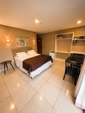 Quarto de hotel com cama, mesa de cabeceira, cadeira e uma pequena escrivaninha com prateleiras.